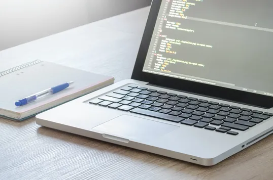 This image contains a closed notebook, a pen, and a laptop in a table.