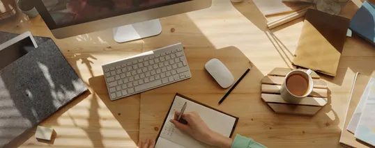 This image contains a person writing in a notebook, a cup of coffee, and a laptop in a table.