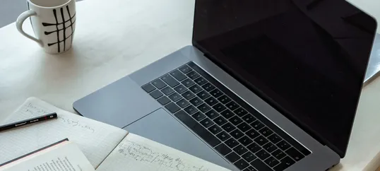 This image contains a notebook with some notes, a cup of coffee, and a laptop in a table.