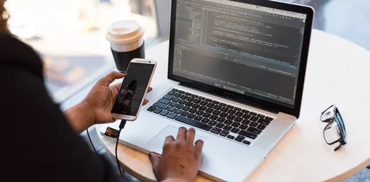 This is a image of a black person holding a mobile, and a laptop with a code editor open.